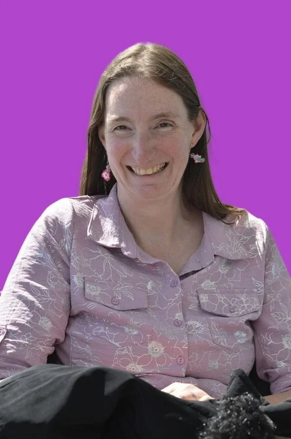 A woman with long brown hair, wearing a light purple embroidered shirt and pink floral earrings, smiling at the camera against a purple background.