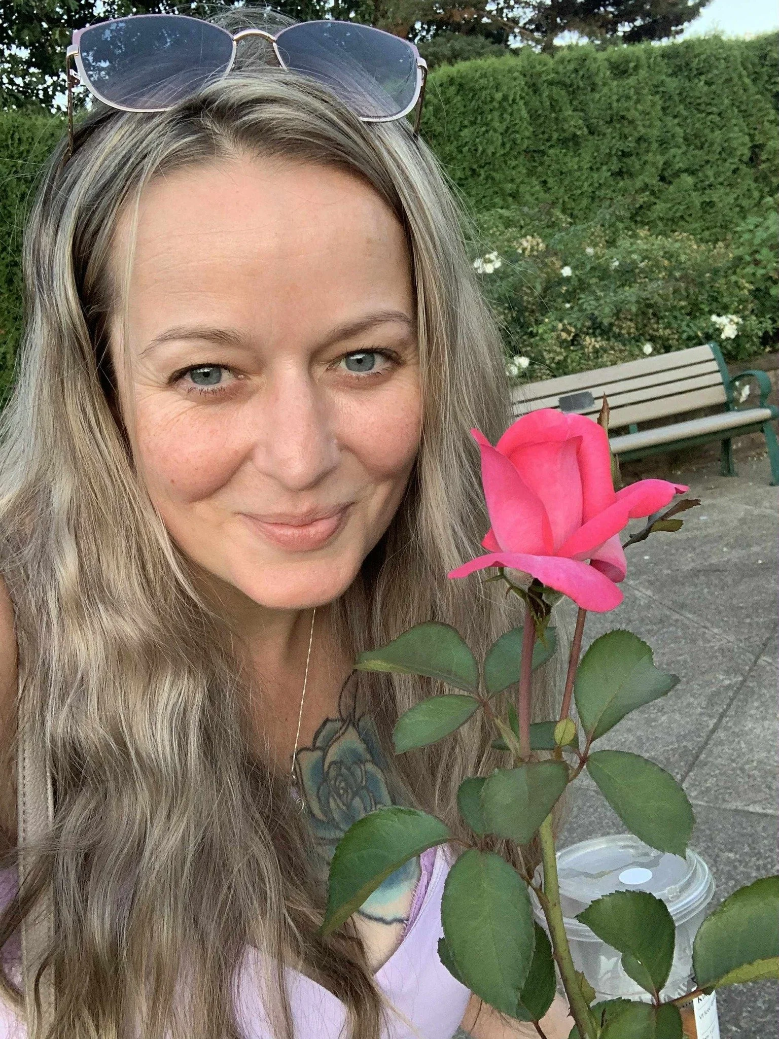 A woman with long wavy blonde hair, blue eyes, and sunglasses on her head taking a selfie outdoors. She is holding a pink rose with green leaves in front of her, and there is a coffee cup visible in the background. The scene is set in a garden or park with greenery and a bench behind her.