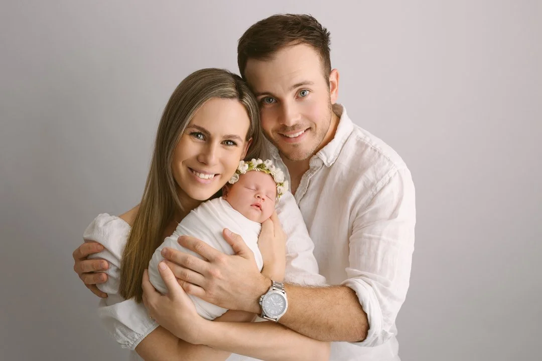 A happy family with a mother, father, and their newborn baby girl posing together against a plain gray background.