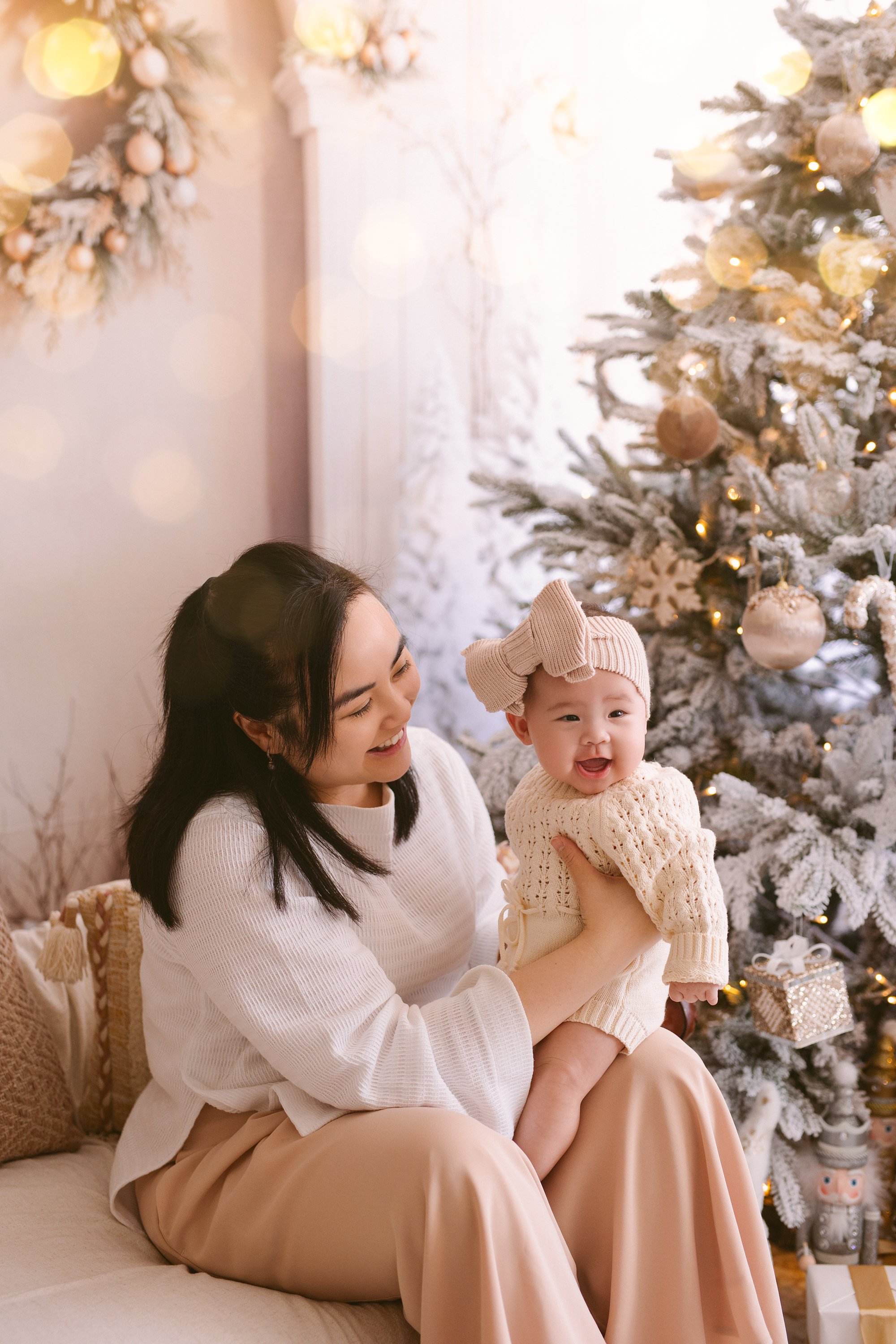 A woman holding a happy baby girl in front of a decorated Christmas tree in a cozy, festive room.
