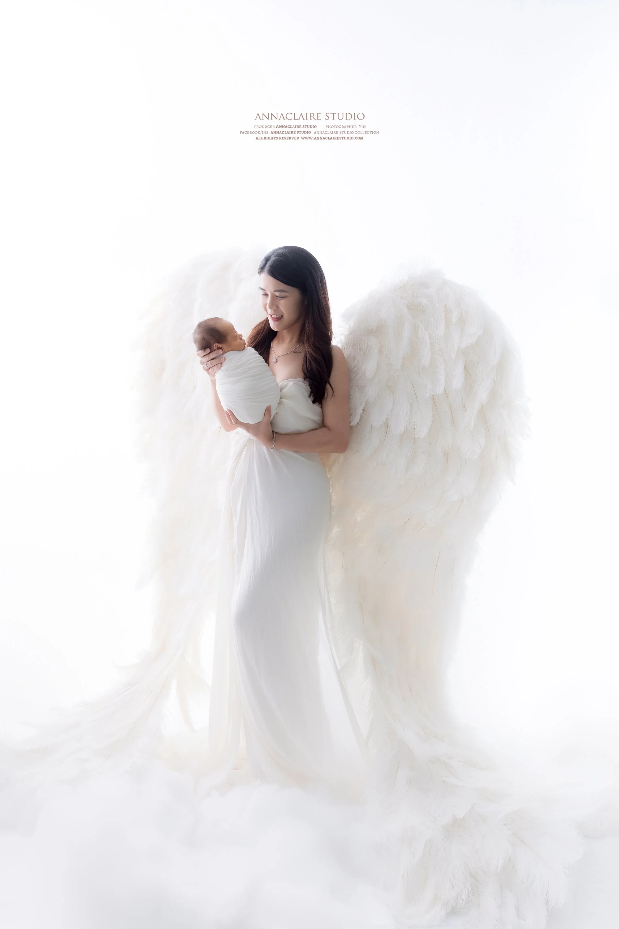 A mum dressed in a white gown holding a swaddled newborn baby, standing with large white feathered angel wings against a white background.