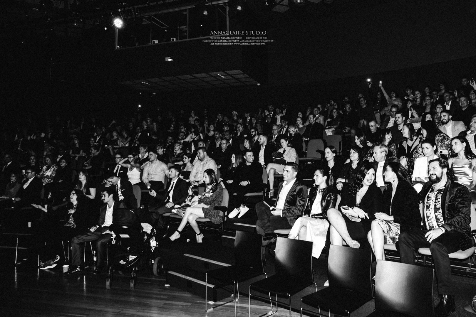 A large audience seated in a dark auditorium, watching a performance or event. The image is in black and white.