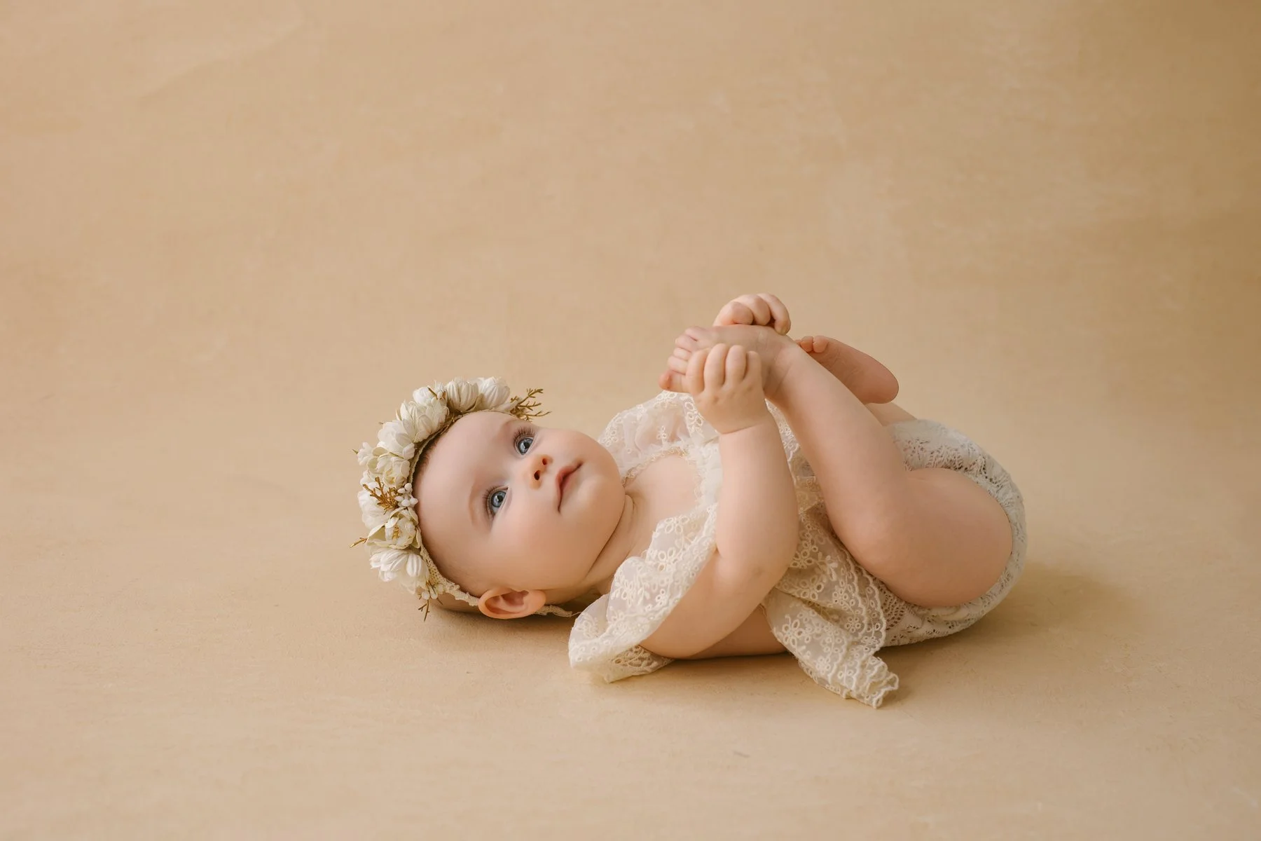 A baby lying on a beige background, wearing a lace dress and a floral headband, holding her feet and looking upward.