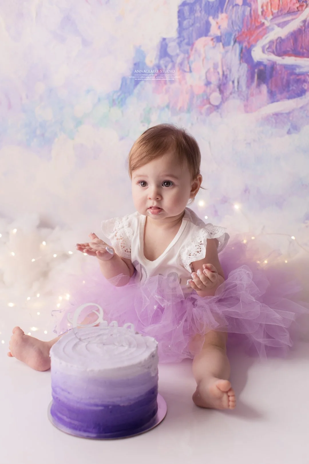 A  one year old girl sitting on the floor in front of a purple gradient cake with a frisbee on top, wearing a white lace top and a purple tulle tutu, with a colorful abstract background and fairy lights.
