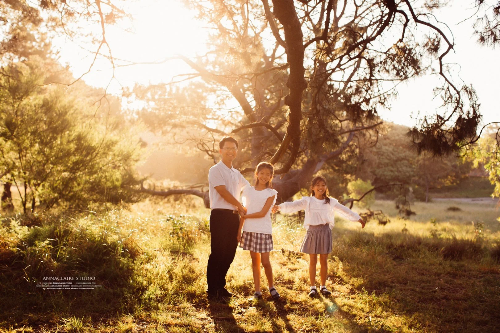 A man and two young girls stand outdoors in in centennial PARK .  during sunset, holding hands, surrounded by trees and grass.