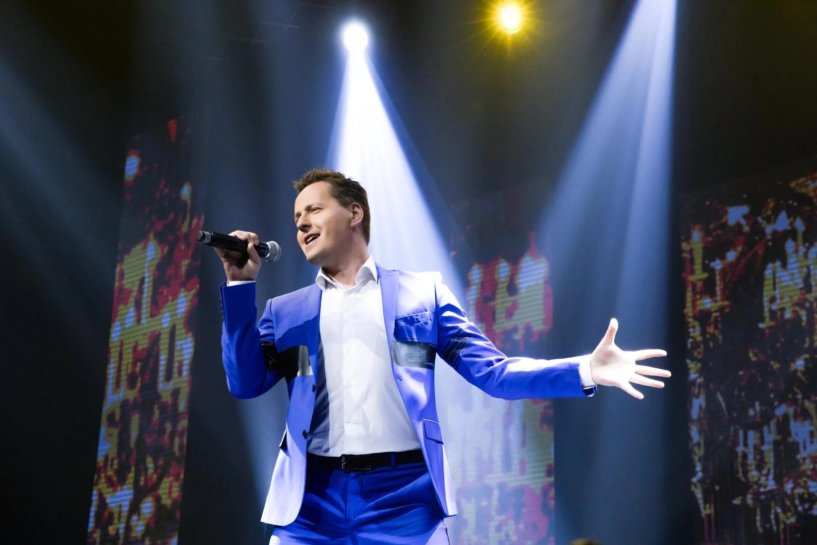 A man in a blue suit singing into a microphone on stage with colorful lights and digital screens in the background.