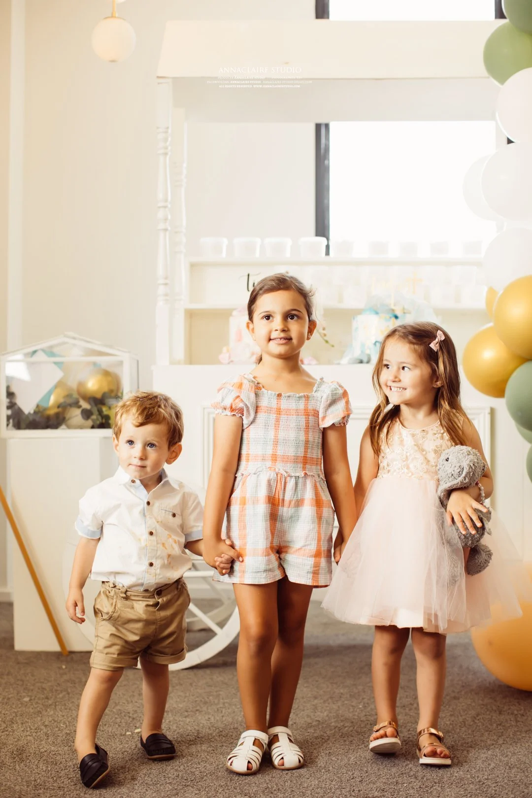 Three young children, two girls and one boy, holding hands at a birthday party with balloons in the background.