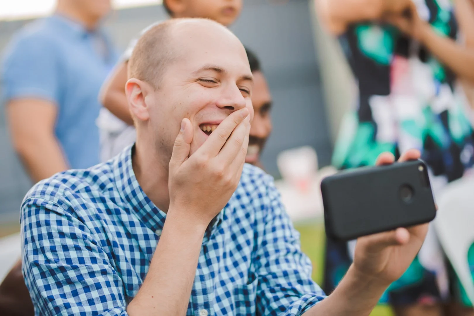 A young man with a bald head and checkered shirt laughs while looking at his phone, surrounded by people at an outdoor gathering.