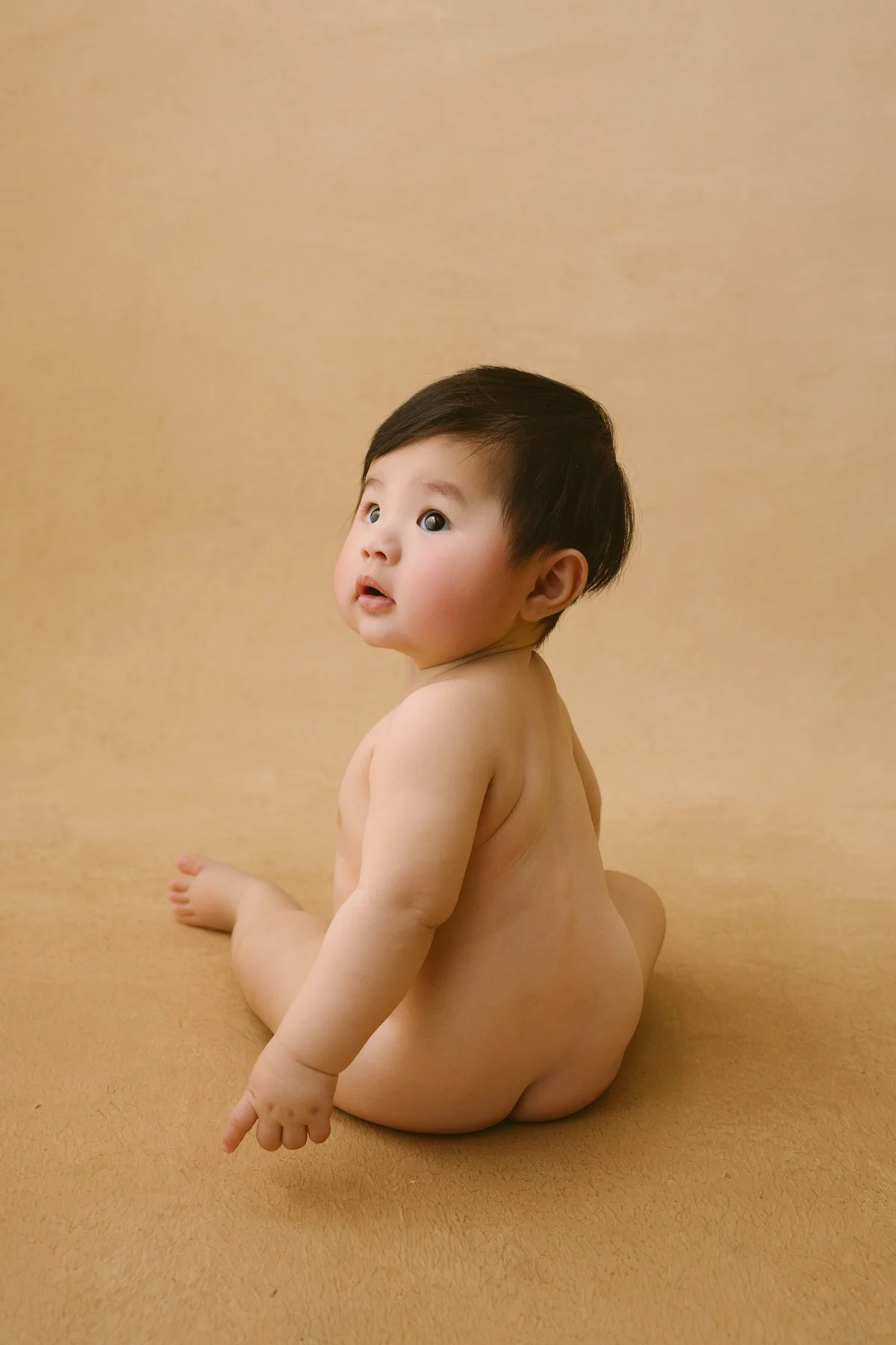 A young child with dark hair, sitting nude on a beige surface and looking up with a curious expression.