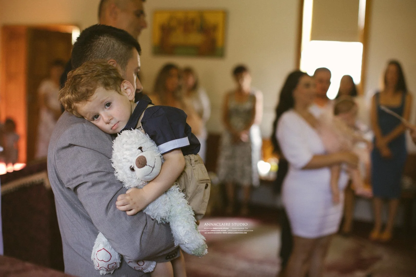 A young boy with curly hair peacefully resting his head on a man's shoulder while holding a white teddy bear, in a gathering room with several women in the background.