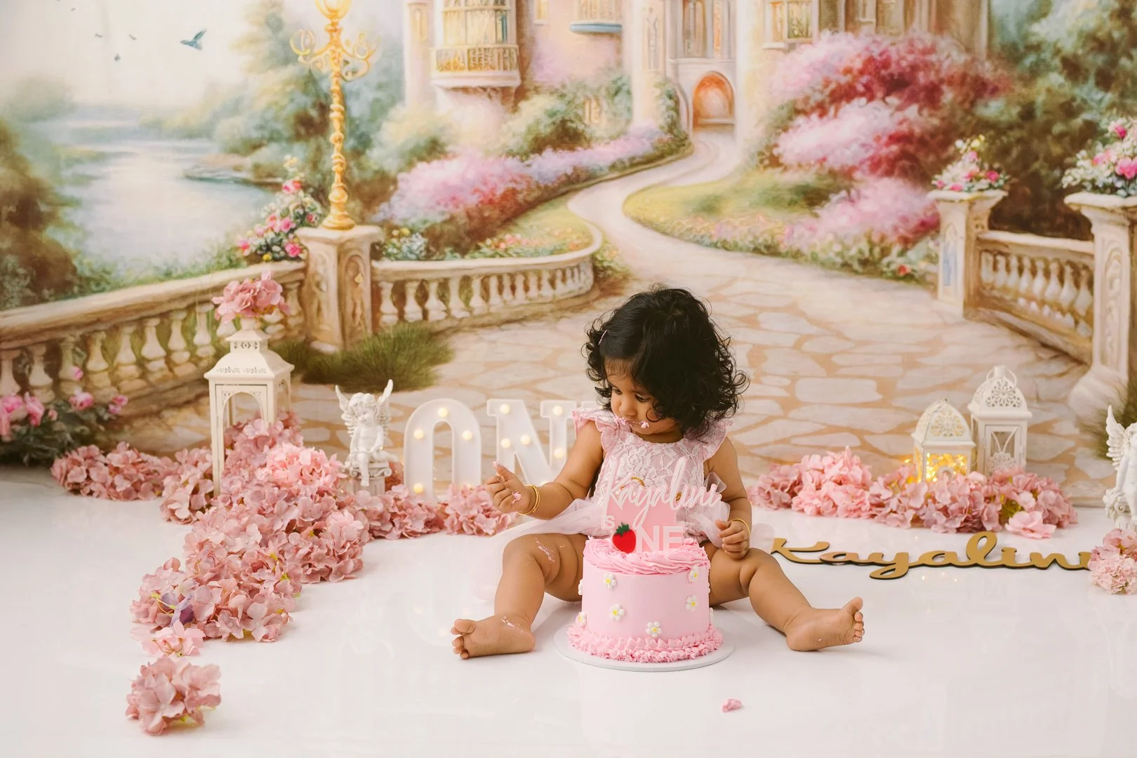 sits on the floor celebrating her first birthday with a pink cake decorated with small white flowers and a pink topper reading 'ONE' and 'Kayalyn'. She is wearing a pink dress and holding a small object, possibly a toy or a candle. The background fea