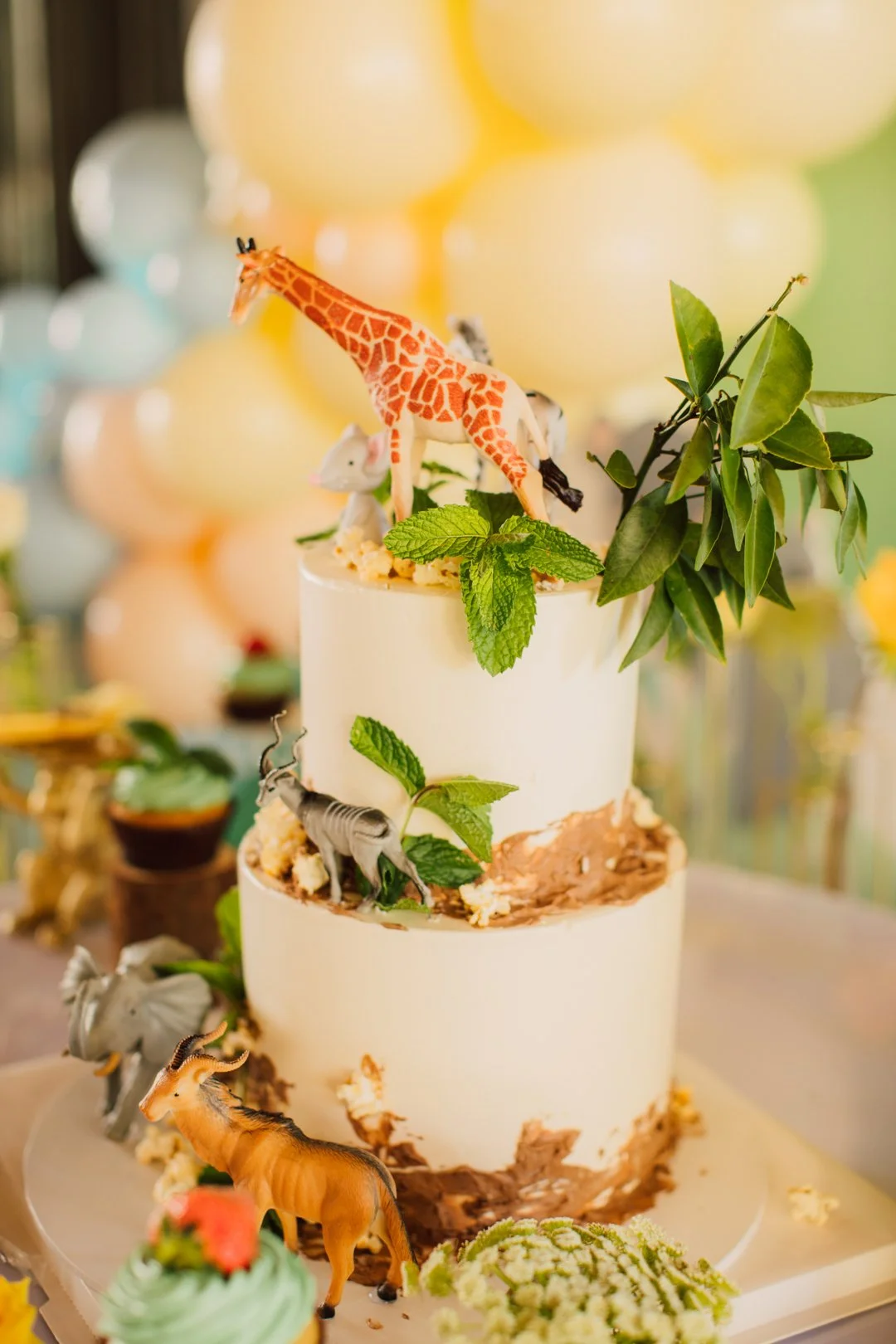 A two-tiered cake decorated with toy safari animals, green leaves, and mint leaves, with pastel-colored balloon decorations in the background.