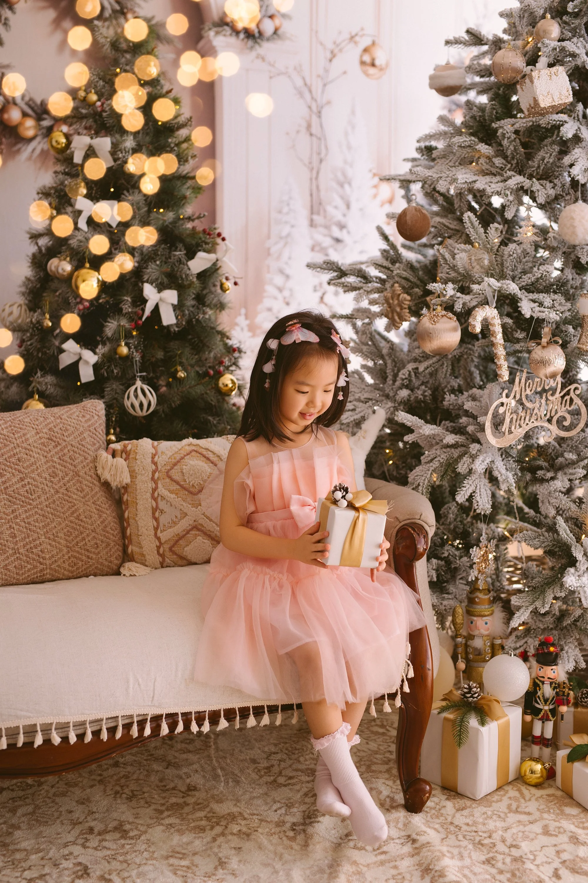 A young girl in a pink dress sitting on a vintage sofa, holding a Christmas gift. She is surrounded by decorated Christmas trees with ornaments, lights, and a "Merry Christmas" sign, with other presents and holiday decorations nearby.