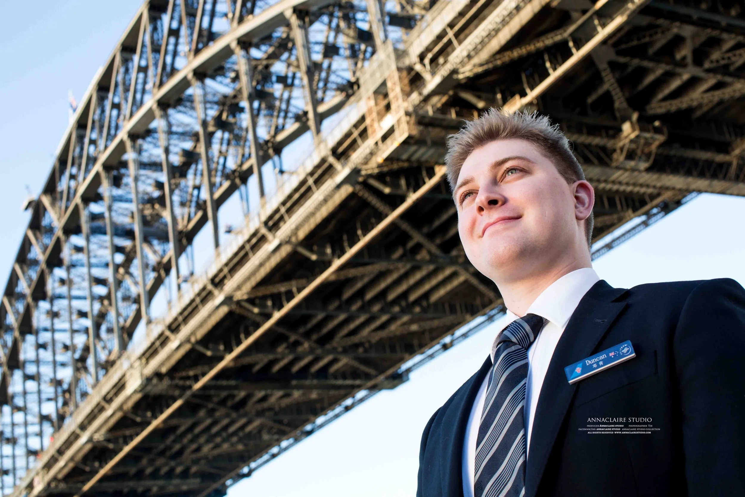 A young china southern airlines male flight attendant in a suit with a name tag stands in front of the Sydney Harbour Bridge, smiling and looking off into the distance.