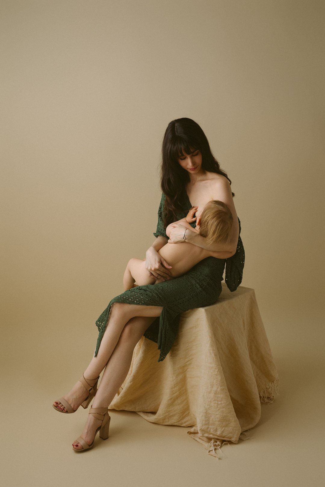 A beautiful mum with long dark hair and in high heels sits on a beige cloth-covered stool, breastfeeding a toddler.
