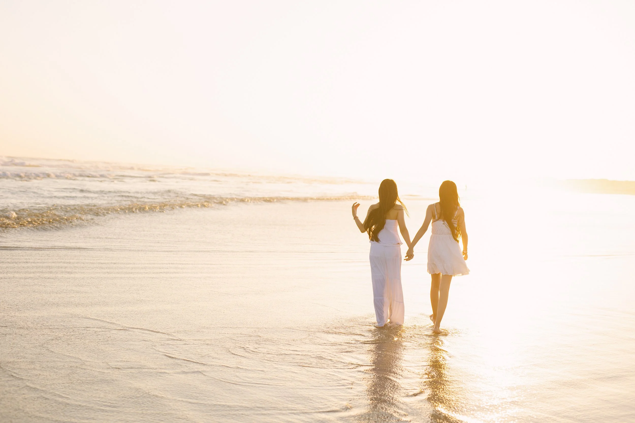 Two women walking hand in hand along a beach at sunset, wearing white dresses with the ocean waves and golden sky in the background.