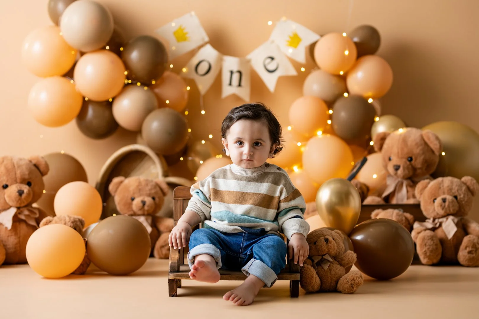 A young child with dark hair and light skin sitting on a small wooden chair, surrounded by teddy bears, balloons, and a 'One' birthday banner. The cake smash photo background features a decorative balloon arrangement and lights.