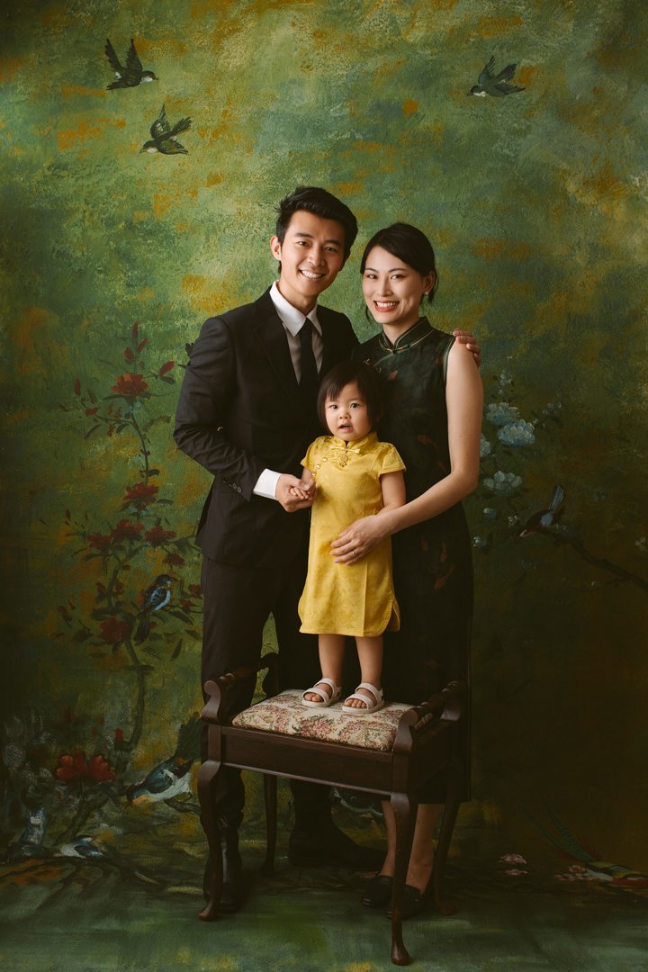 A family of three posing for a portrait with a painted nature background. The father and mother stand behind a small table, with the father holding the child's hand. The child, dressed in a yellow dress, stands on the table. The background features g