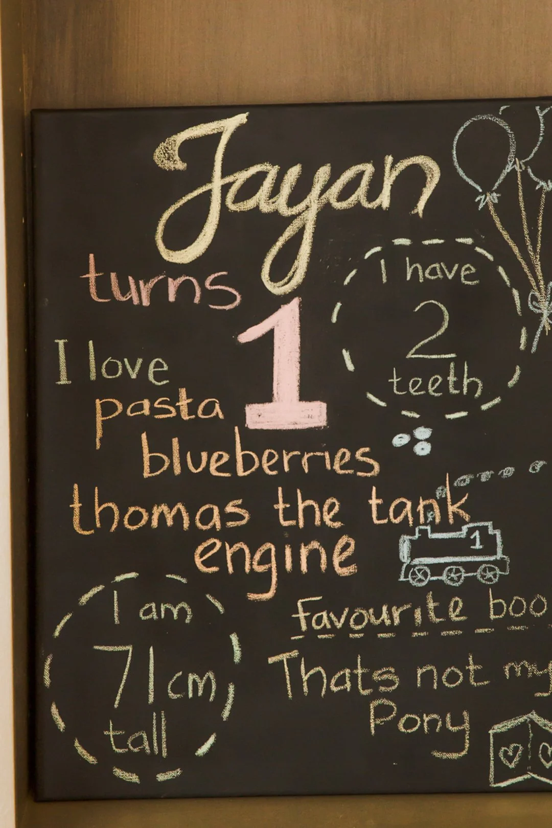 Decorative chalkboard with colorful handwritten text and drawings celebrating a child's first birthday. The message includes phrases like 'Jaylan turns 1', 'I love pasta', 'blueberries', 'Thomas the tank engine', and 'I am 71 cm tall'. There are dood