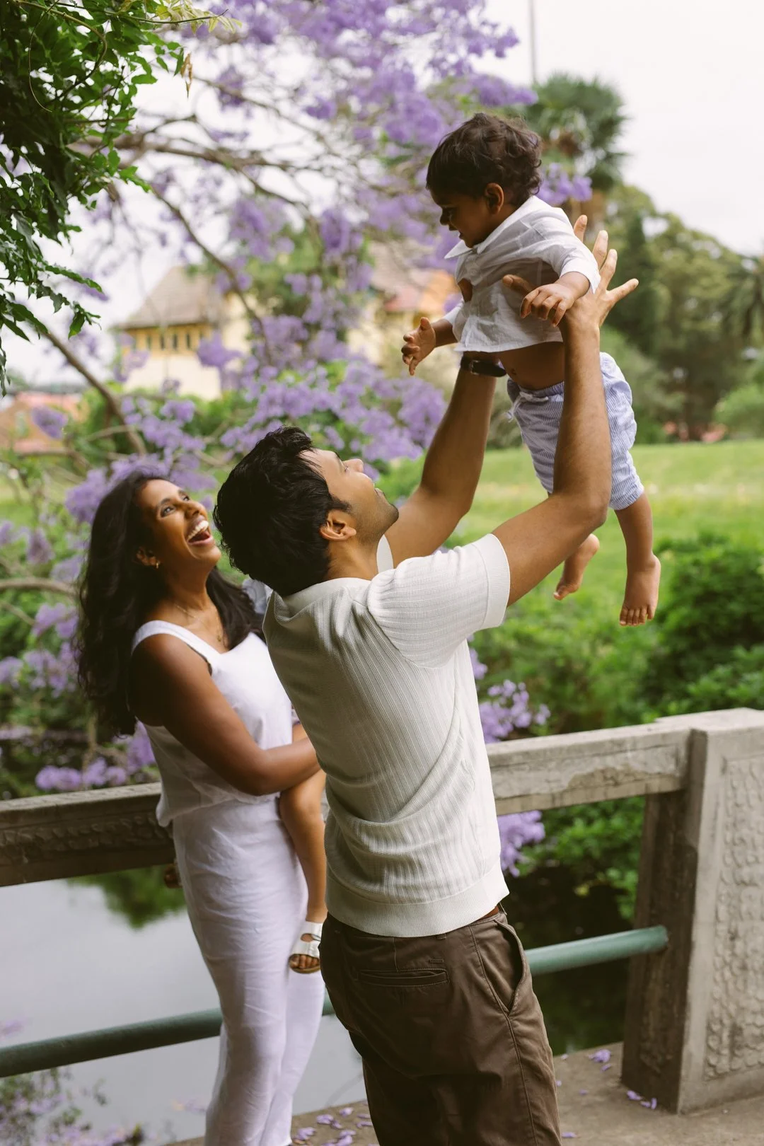 A joyful family enjoying time outdoors near a pond, with a man lifting a young boy in the air, a woman smiling in the background, surrounded by purple flowering trees and greenery.