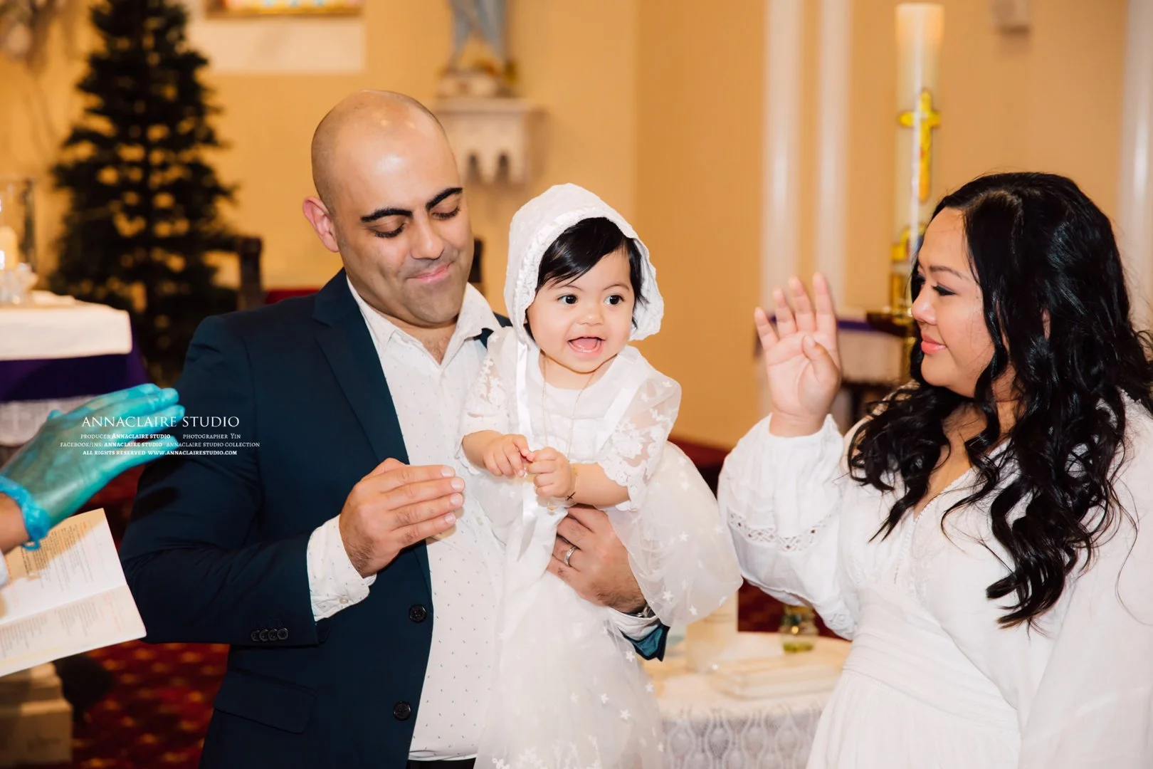 A family celebrating a religious event, with a man holding a young girl dressed in white, and a woman raising her hand for a blessing or prayer, in a church decorated with candles and Christmas trees.