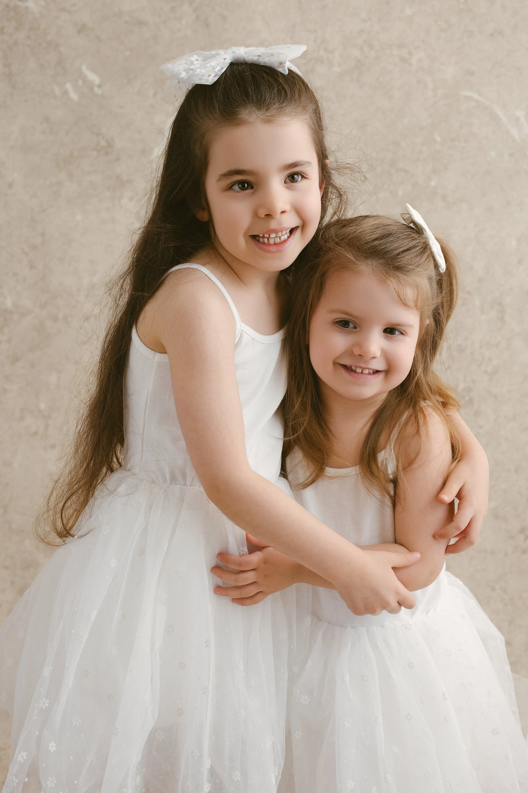 Two young girls in white dresses hugging and smiling, standing in front of a beige textured background.