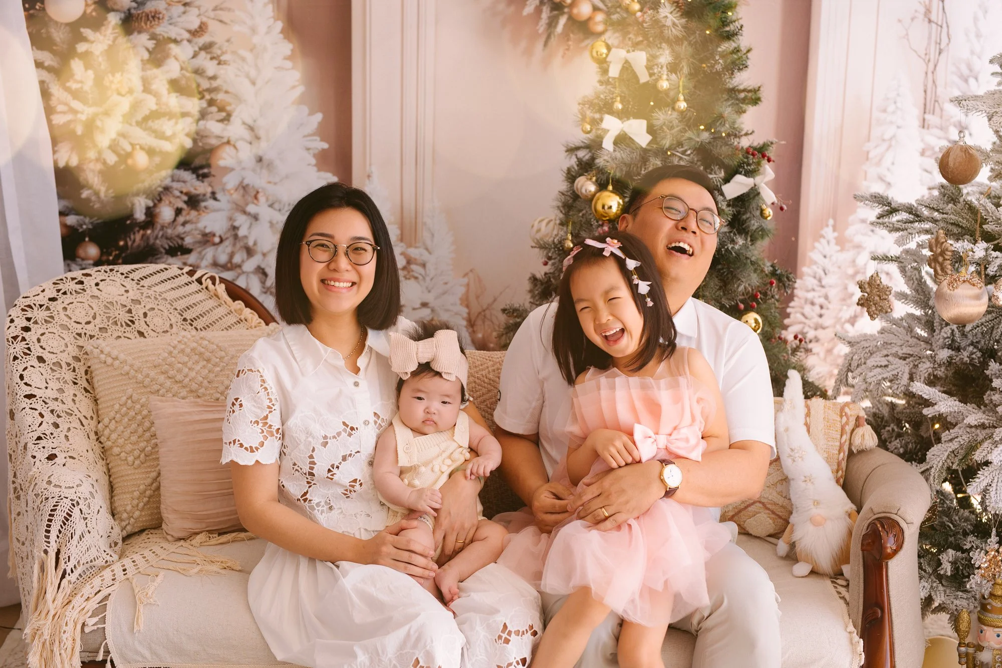 A family of four sitting on a beige couch with Christmas trees and decorations in the background, smiling and laughing.