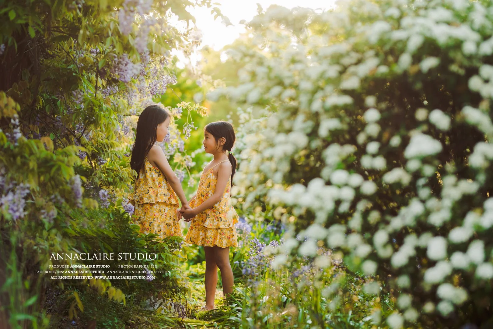 Anna AND Claire  in yellow floral dresses holding hands and facing each other in a lush garden with blooming flowers and greenery, sunlight shining through the trees.