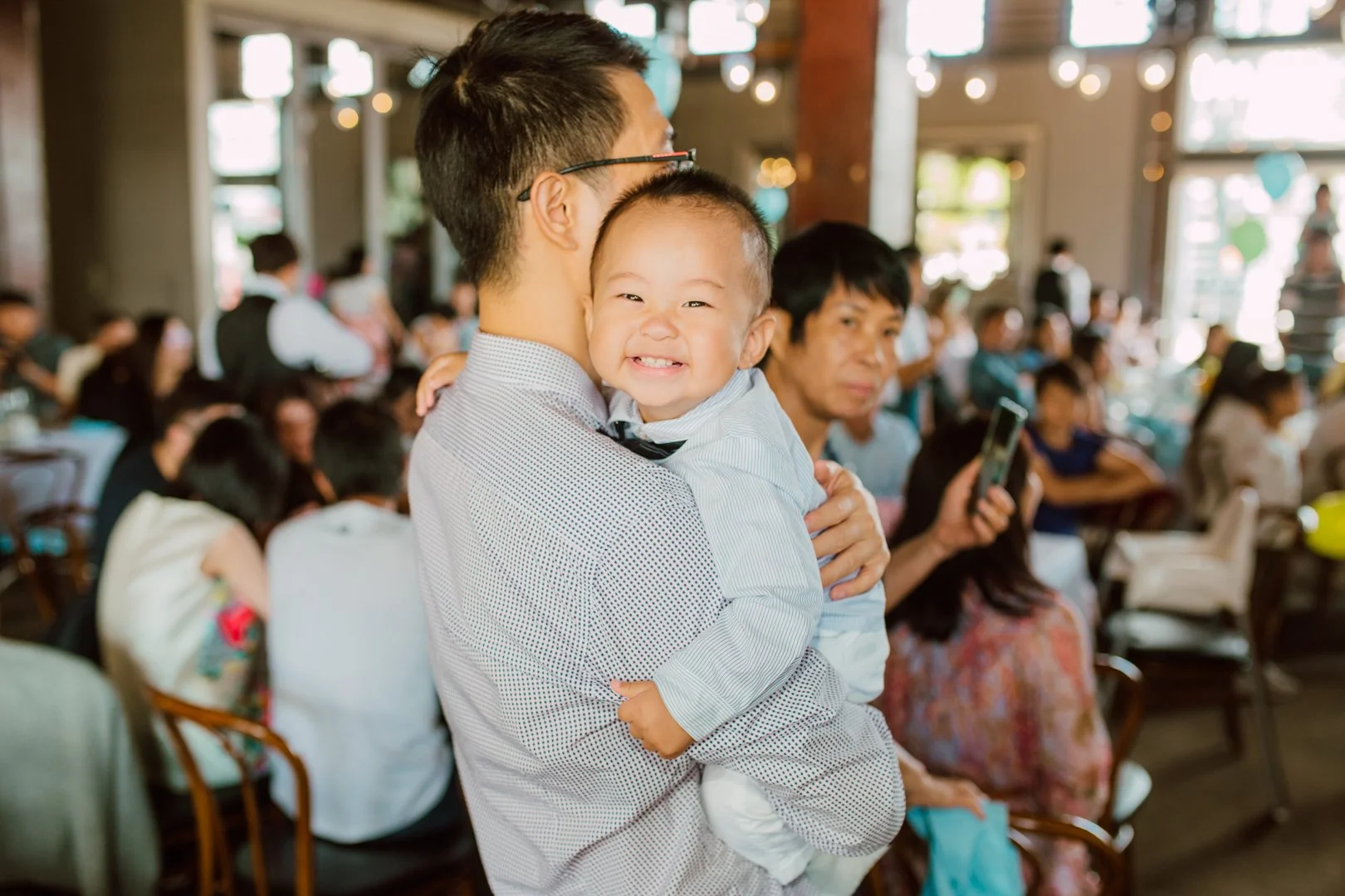 A man in glasses holding a smiling young boy in a white shirt at a crowded indoor event.