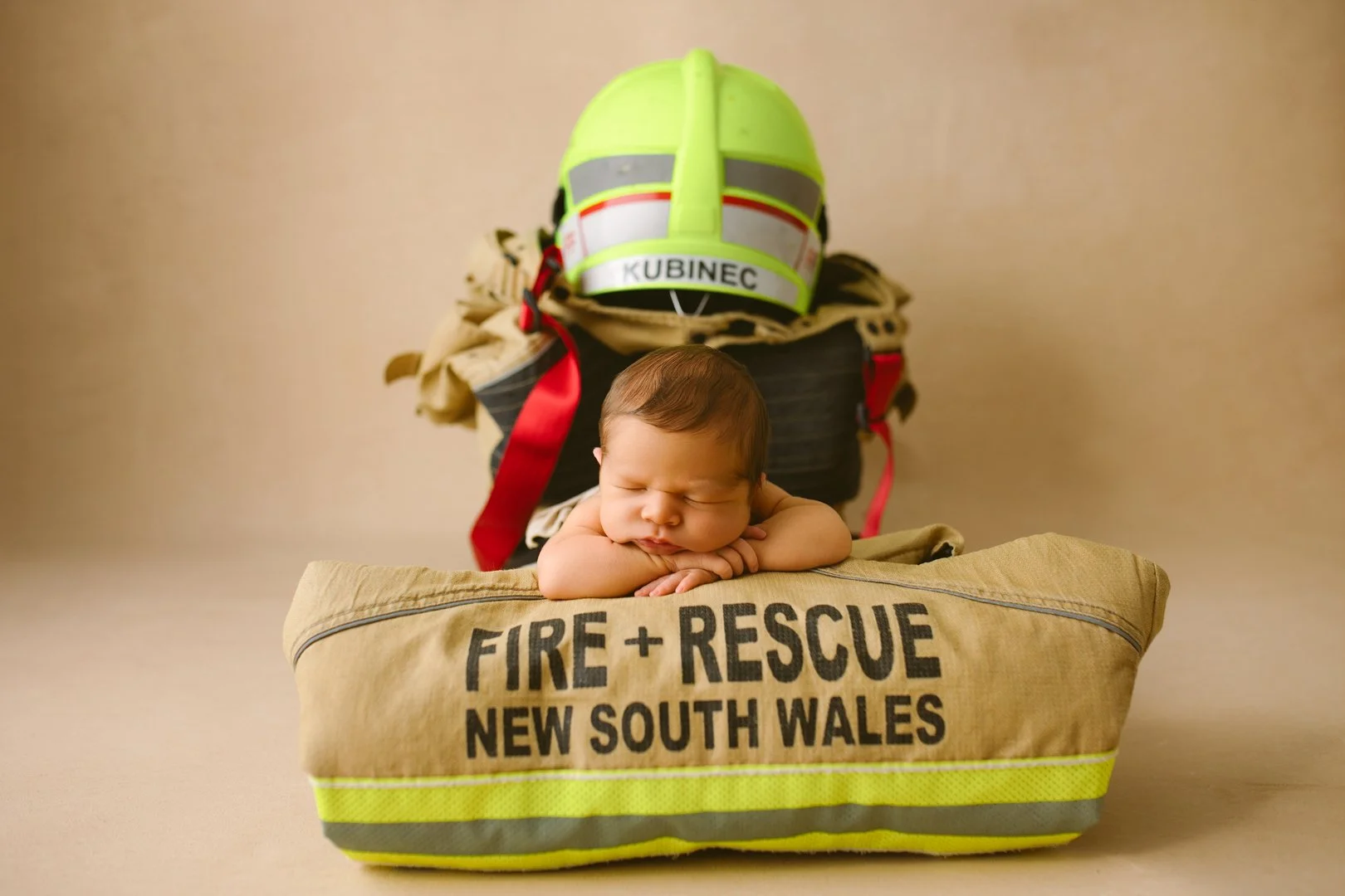 A newborn baby sleeping on a beige fire rescue bag with the words "FIRE + RESCUE NEW SOUTH WALES" printed on it, with a firefighter helmet labeled "KUBINEC" and fire gear in the background.