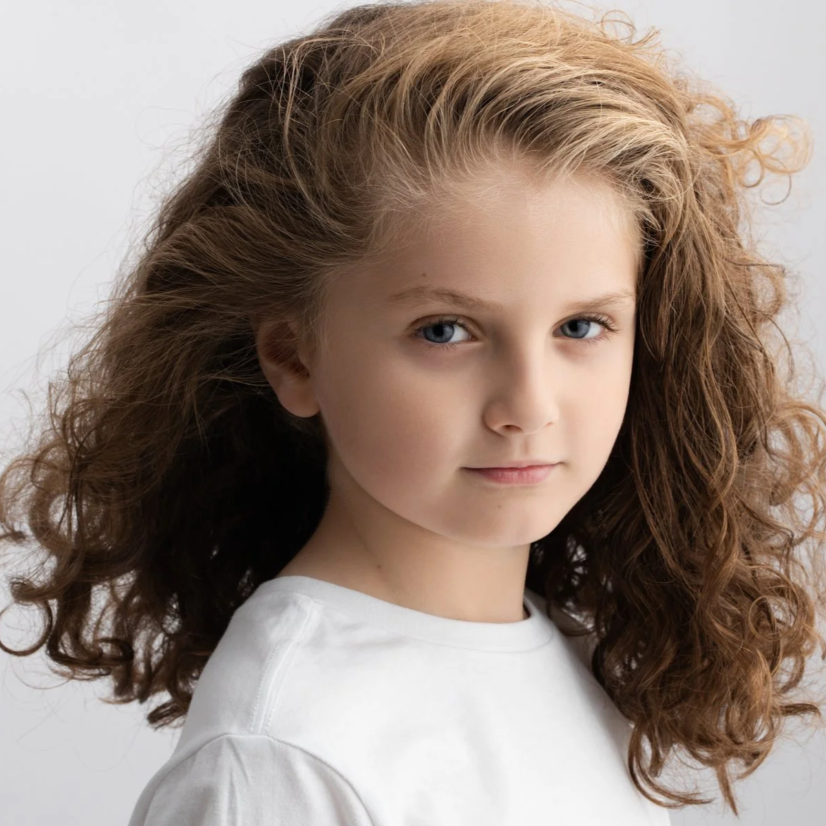 Close-up portrait of a young girl with curly, light brown hair and blue eyes, wearing a white shirt against a plain background.