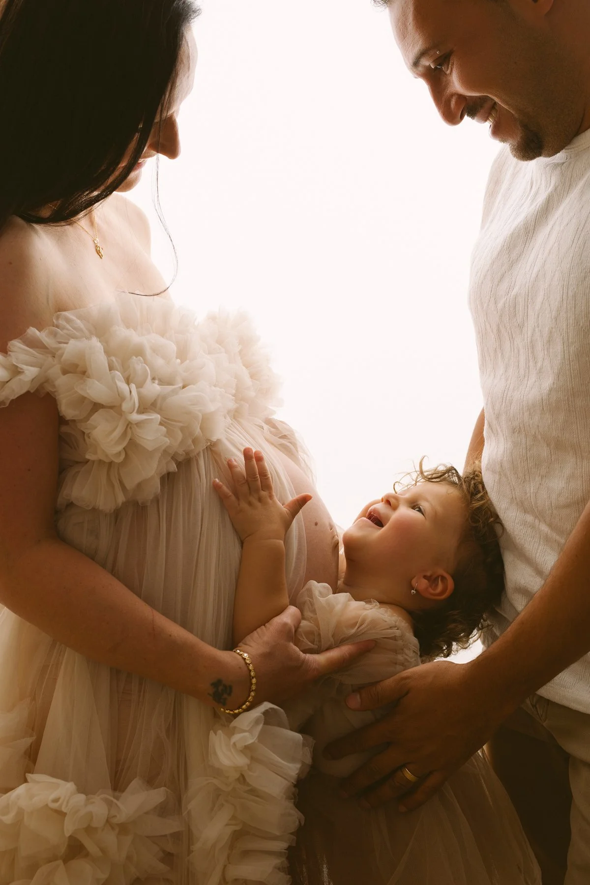 A pregnant woman with a floral dress touching her belly, a smiling toddler girl lying on her mother's belly with an adult man leaning in close, all looking at each other affectionately.