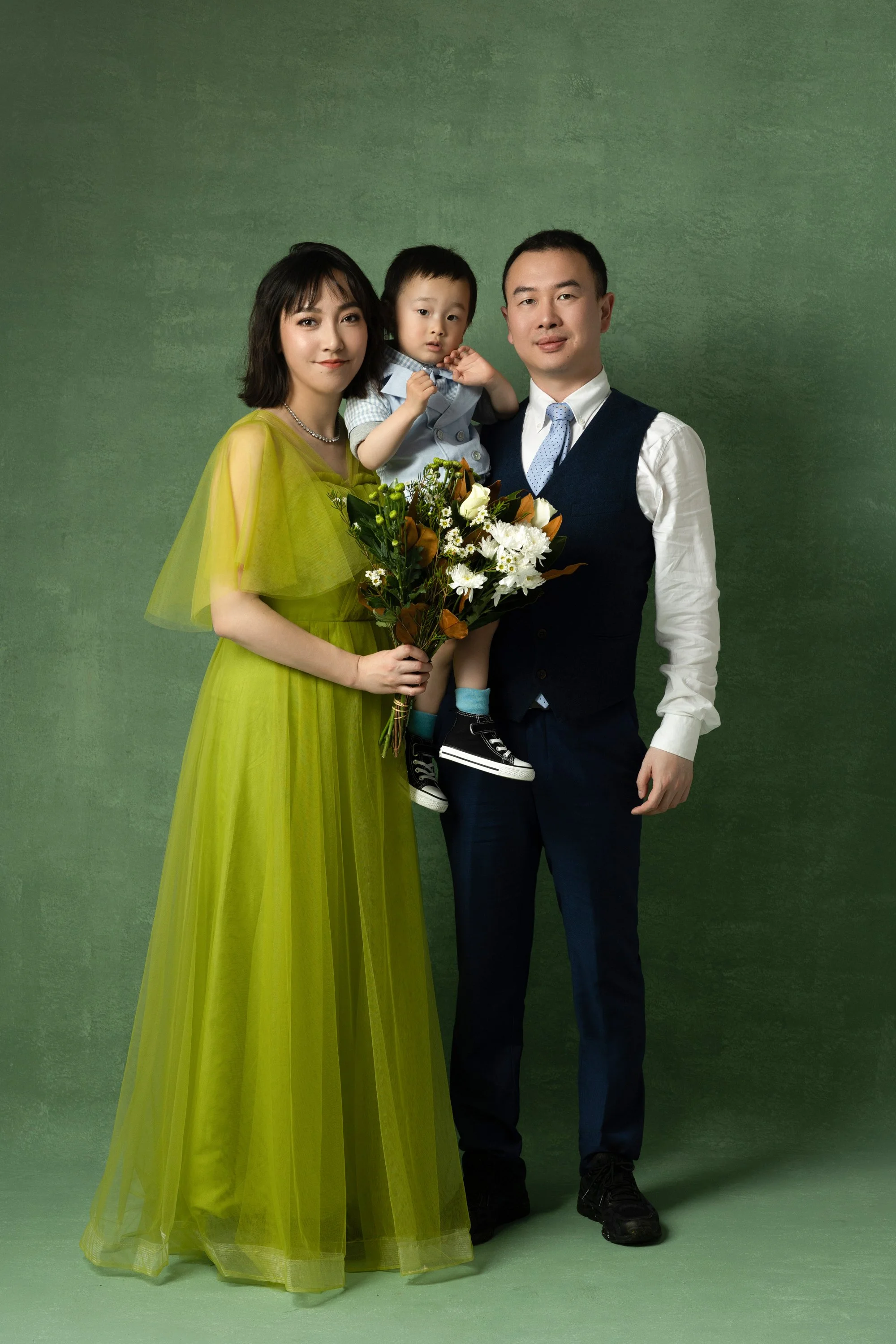 A family of three posing together, the woman holding a bouquet of flowers, the man holding a young boy, all standing against a green background.