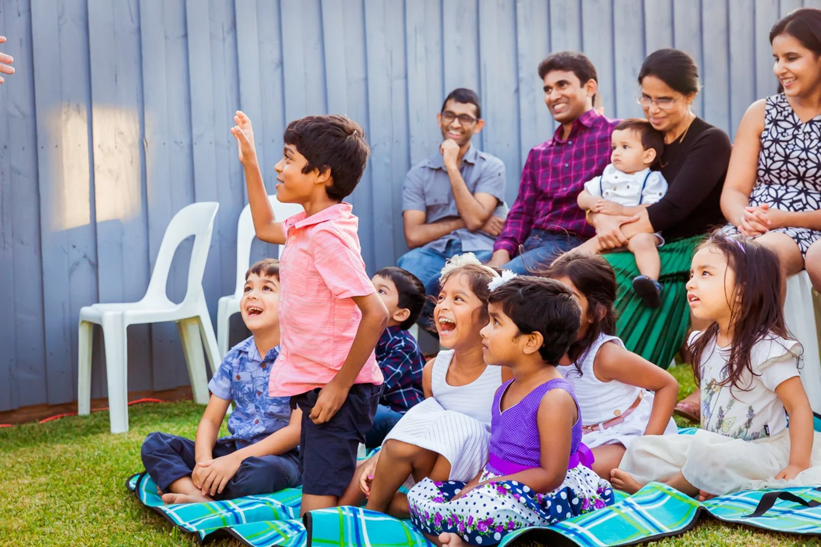 Children sitting on a blanket and an adult sitting on white plastic chairs, watching and smiling, during an outdoor gathering.