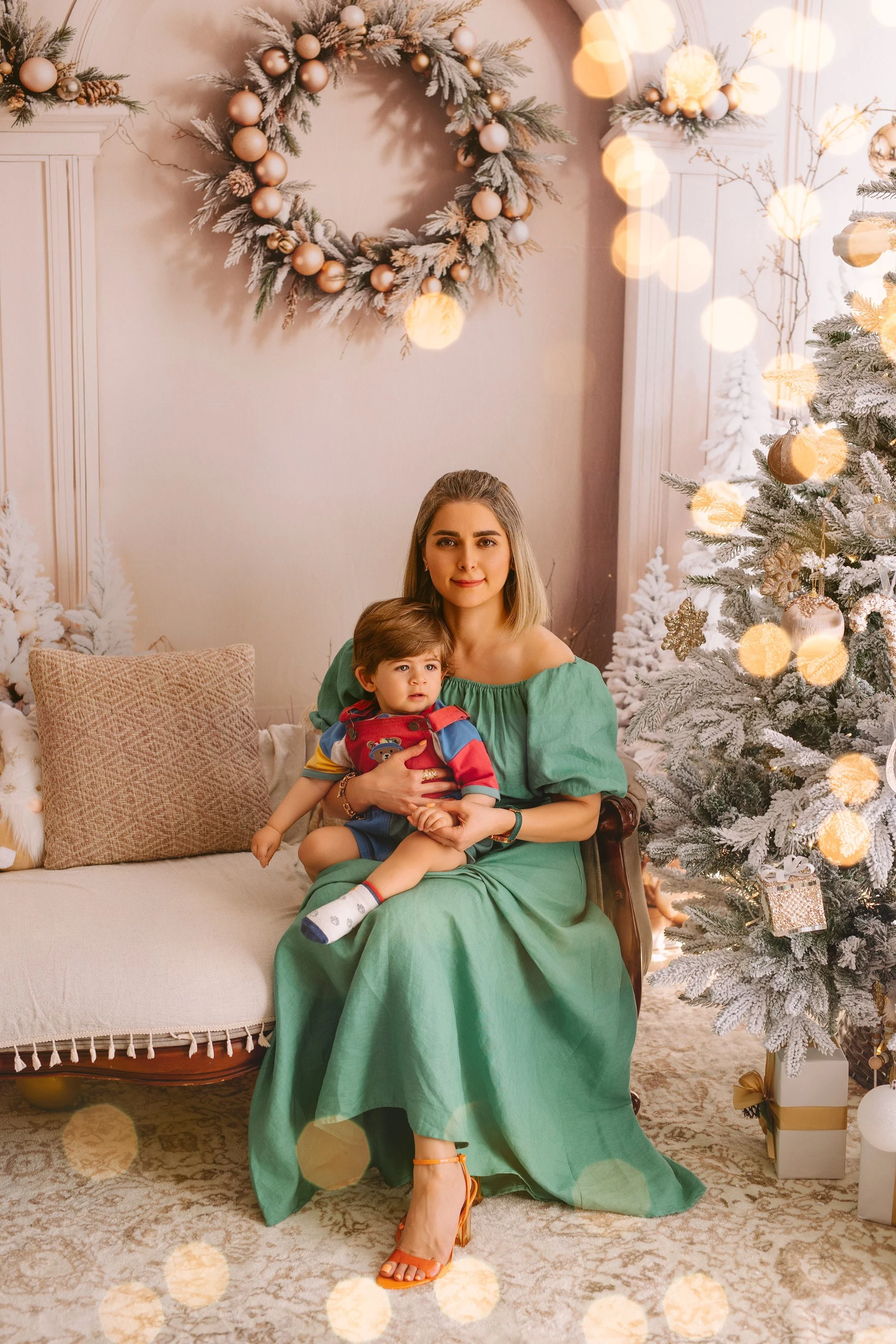 A mum in a green dress sitting on a vintage sofa, holding a young boy in a colorful hoodie, in a Christmas decorated room with a snow-flocked Christmas tree and a wreath on the wall.