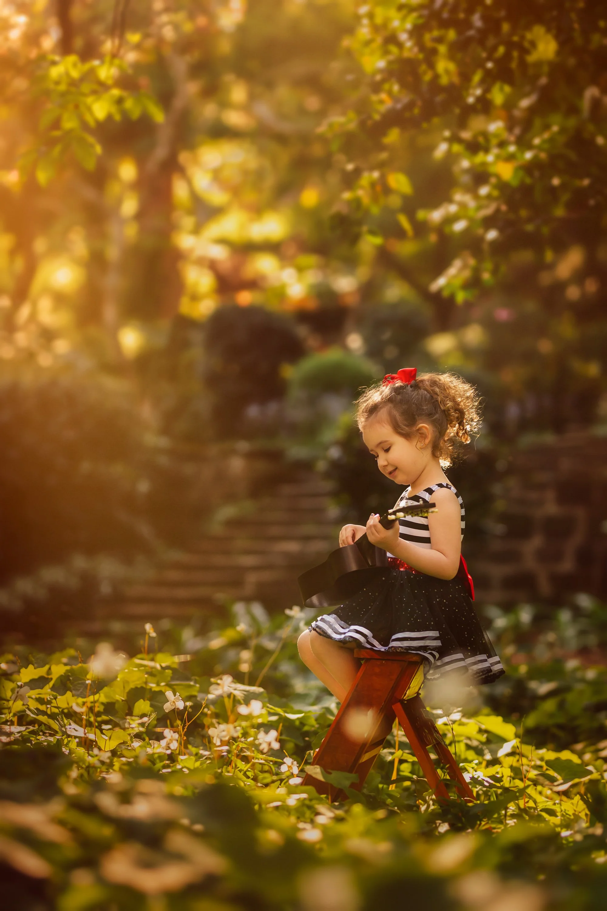 A young girl with curly hair, wearing a black and white striped top, a black skirt, and a red bow, sitting on a small wooden chair outdoors, playing an acoustic guitar among green foliage and white flowers during golden hour.
