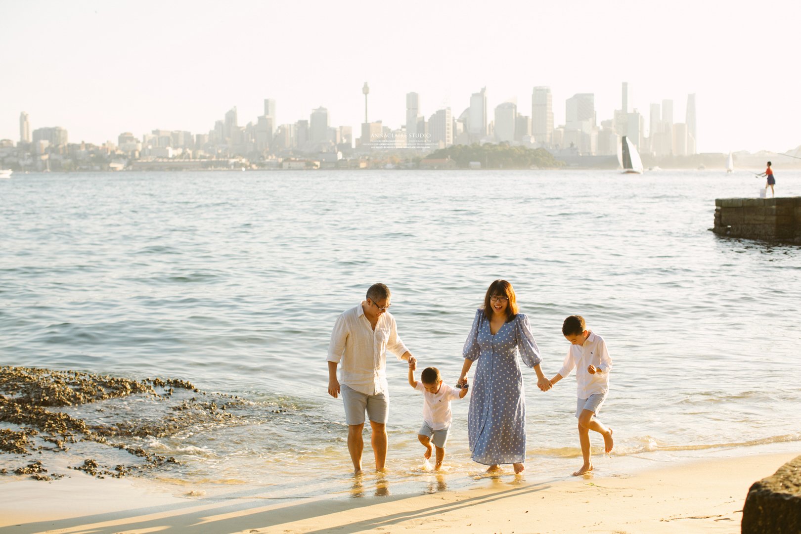 Family of four holding hands and playing in the water on a beach with a city skyline in the background.
