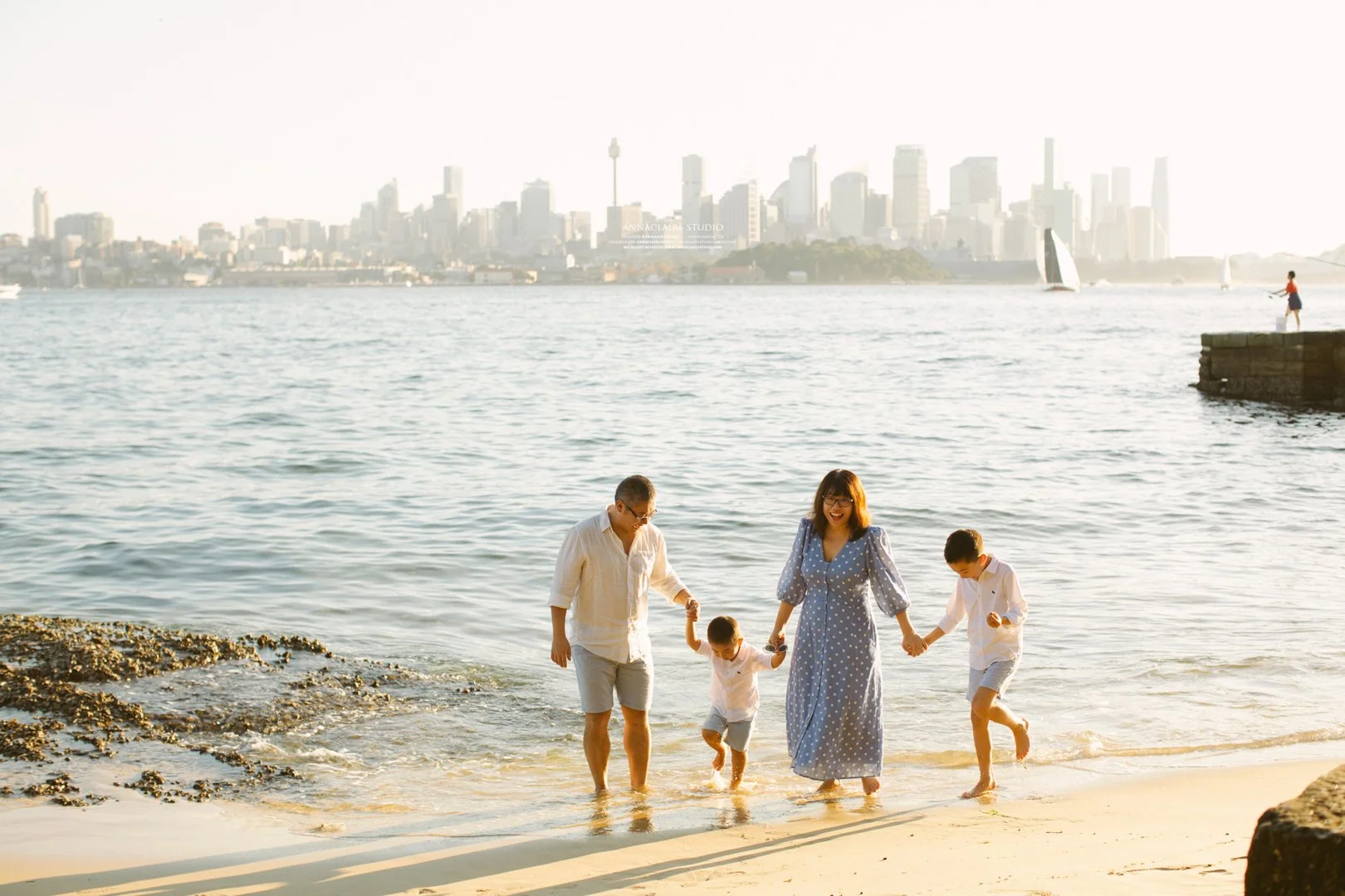 A family of four walking along the beach near water, with a city skyline in the background. The family includes two adults and two children, all dressed in light-colored clothing, holding hands, and enjoying the sunny weather.
