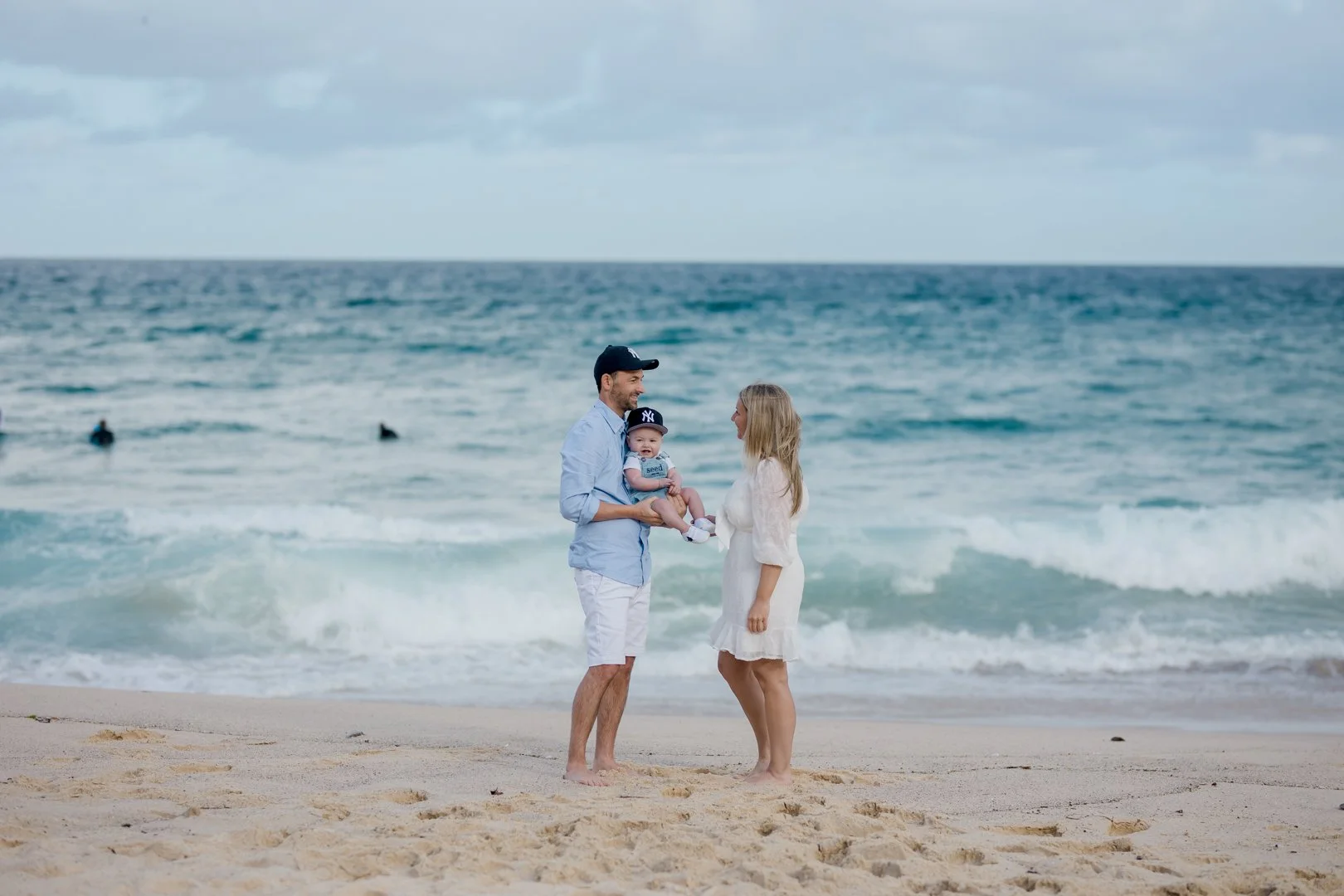 A family of three standing on a sandy beach near the ocean, with the father holding a baby and the mother standing opposite him, engaging in a conversation. The background features waves and a few surfers in the water, under a cloudy sky.