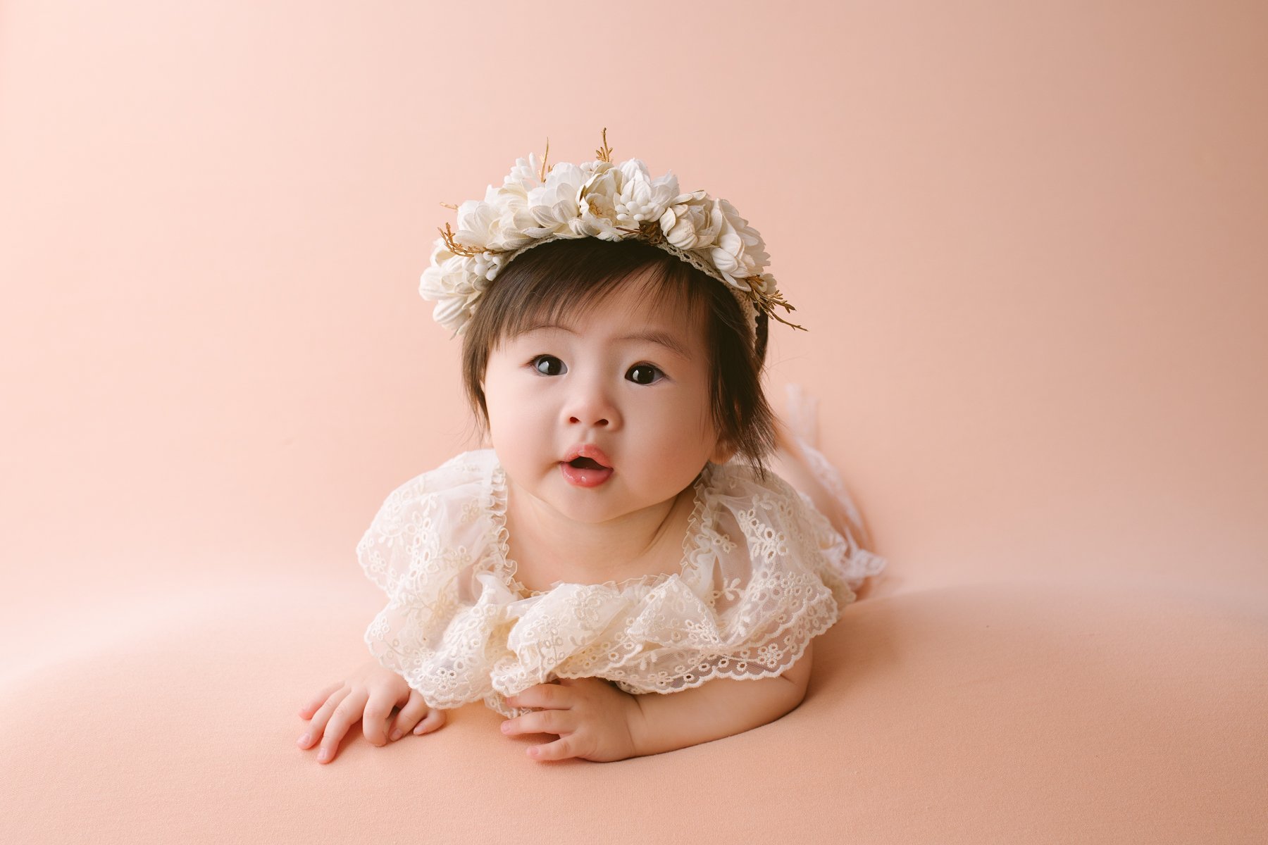 A baby girl lying on a peach-colored surface, wearing a cream lace dress and a floral headband, looking at the camera with a curious expression.