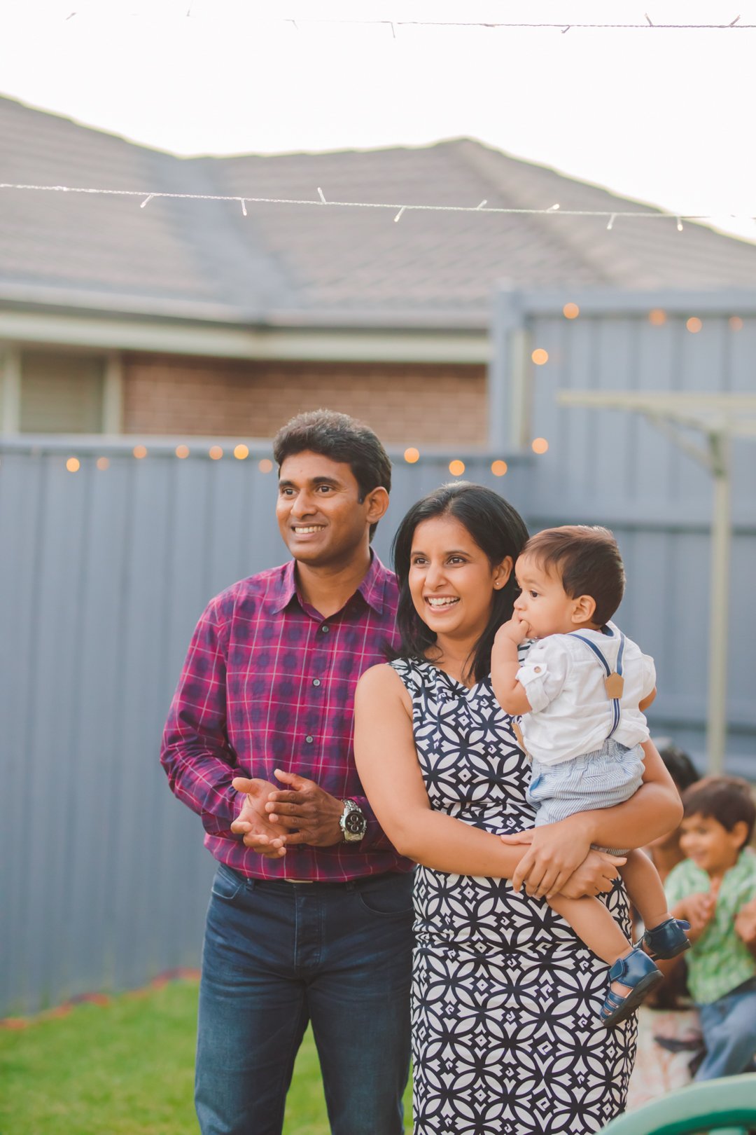 A family gathered outdoors in a backyard with string lights, including a man in a red plaid shirt, a woman in a patterned dress holding a young child, and a boy in a green shirt in the background.