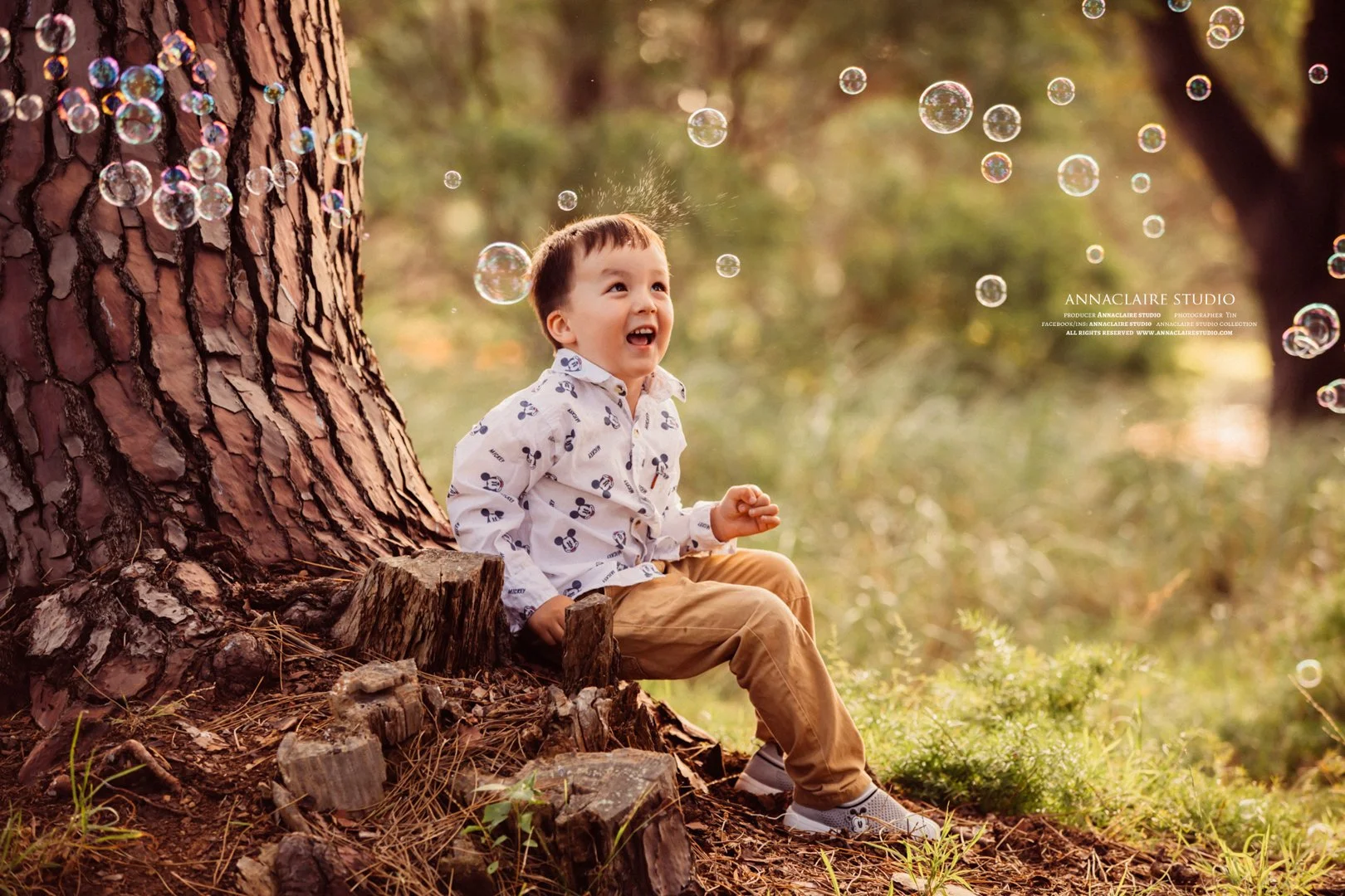 A young boy sitting on the ground next to a large tree trunk with bubbles floating around him in a forest setting, smiling and looking up.