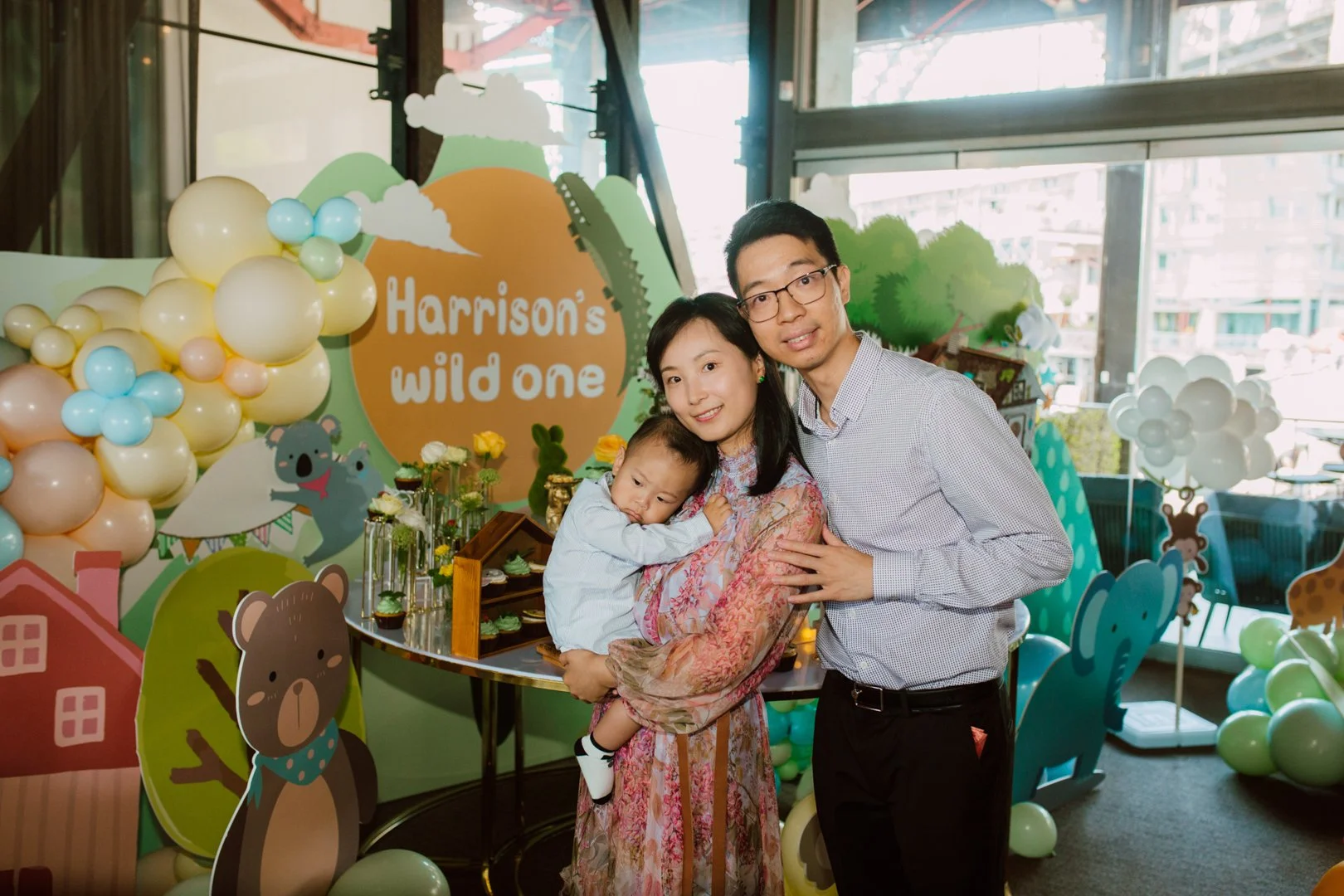 A family of three posing at a jungle-themed birthday party with decorations including balloons and cartoon animal cutouts, with a sign reading 'Harrison's wild one' in the background.