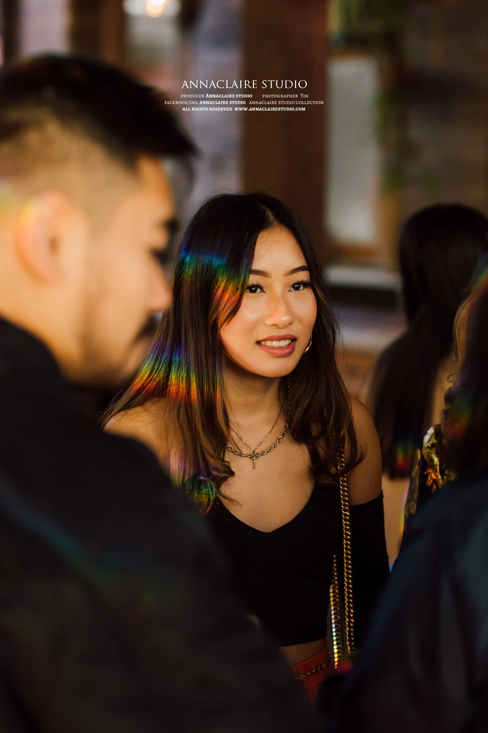 A woman with rainbow-colored streaks in her hair, wearing a black off-shoulder top and gold jewelry, smiling while at a social gathering.