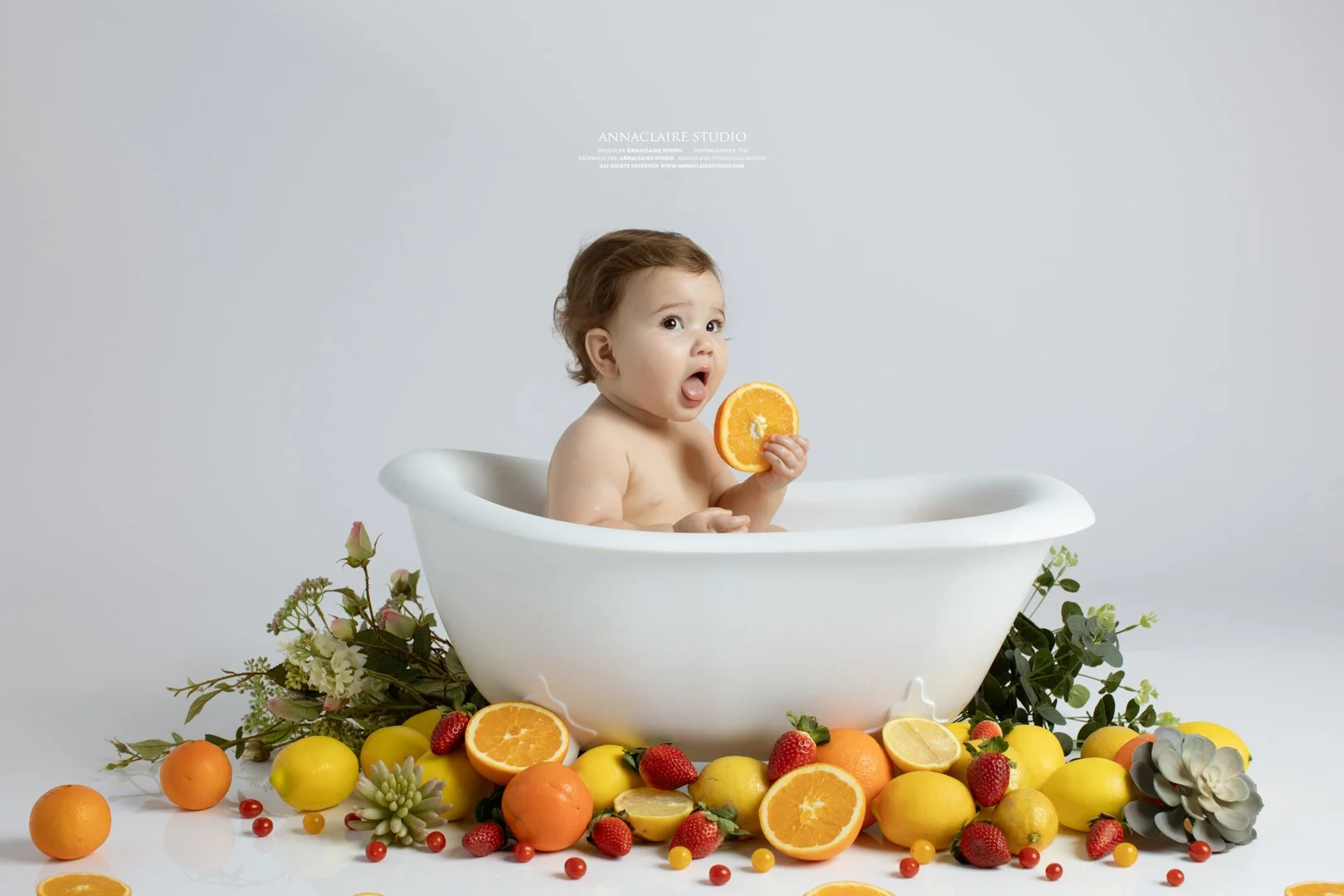 1 yr old baby  sitting in a white bathtub surrounded by an assortment of colorful fruits including oranges, strawberries, lemons, and small red berries, with a plain light-colored bubble bath photo background.
