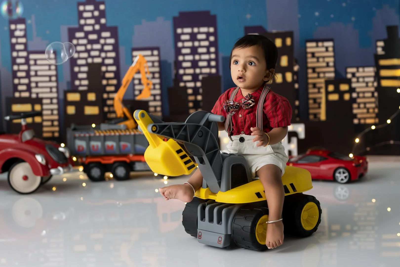A  one year old baby in a red shirt and beige suspenders riding a yellow toy construction excavator in a play area with a cityscape backdrop and toy cars in the background.