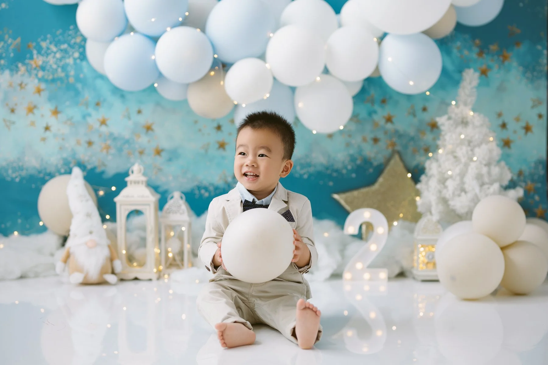 A young boy sits on the floor holding a white ball, surrounded by holiday decorations including white balloons, a Christmas tree, lanterns, a plush gnome, and illuminated stars, with a blue and gold starry background.