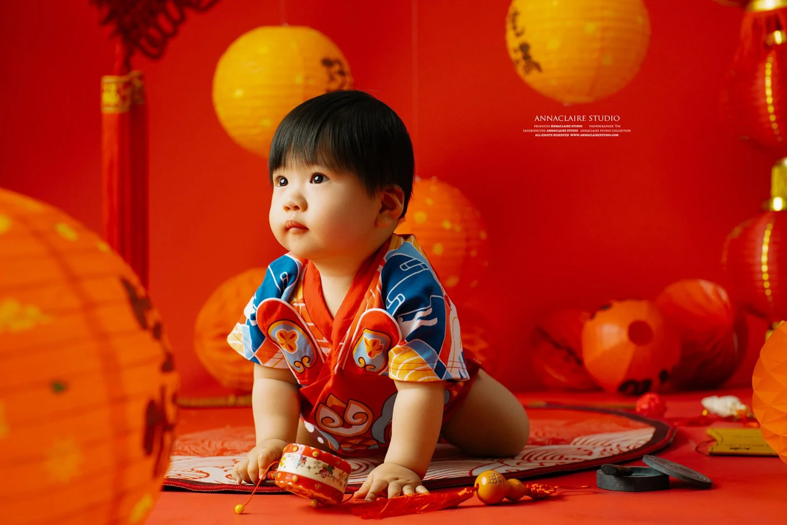 1 year old boy in traditional chinese clothing, surrounded by orange paper lanterns, on a red background, posed on a red mat.