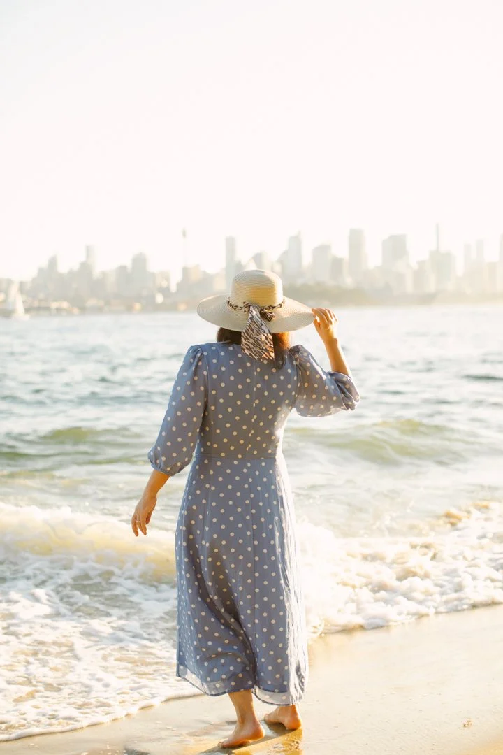 A woman dressed in a blue polka dot dress and wide-brimmed sun hat walks along a sandy beach near the water, with a sydney city skyline in the background.
