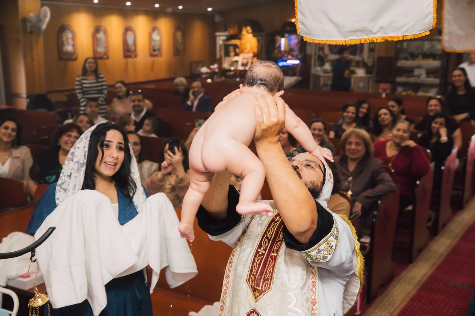 A man holding a baby during a christening ceremony in a church with seated and standing spectators watching and smiling.